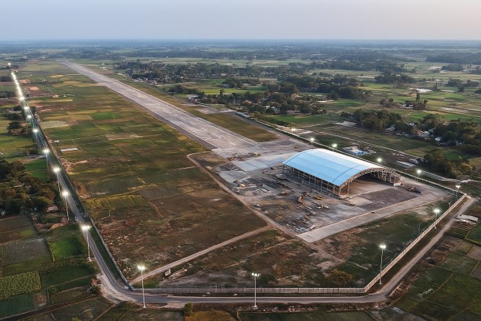 A high-resolution aerial view of the Lalmonirhat airbase in northern Bangladesh, showing the long runway, a large hangar under construction, floodlights along the perimeter, and surrounding green fields.