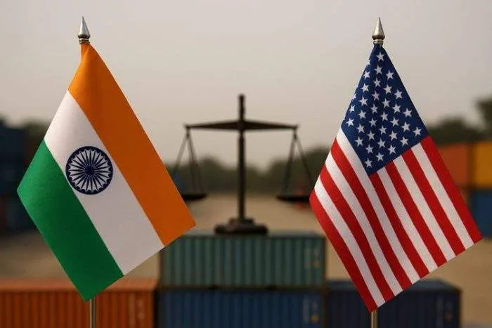 Flags of India and the United States on poles with a balance scale in the background, set against stacked shipping containers.