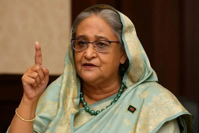 A photograph of Sheikh Hasina, the Prime Minister of Bangladesh, speaking firmly during exile, wearing a light green saree with a national flag pin.
