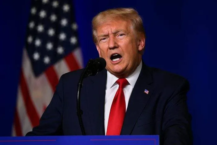 U.S. President Donald Trump speaking at a podium with the American flag blurred in the background.