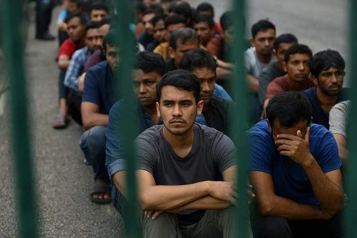 A group of detained South Asian men sit on the ground behind green metal bars, looking weary and anxious.