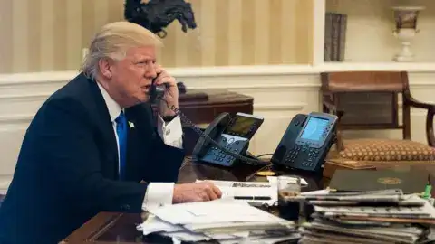 President Donald Trump speaks on the phone in the Oval Office with American flags behind him, symbolizing diplomacy and global discussions.