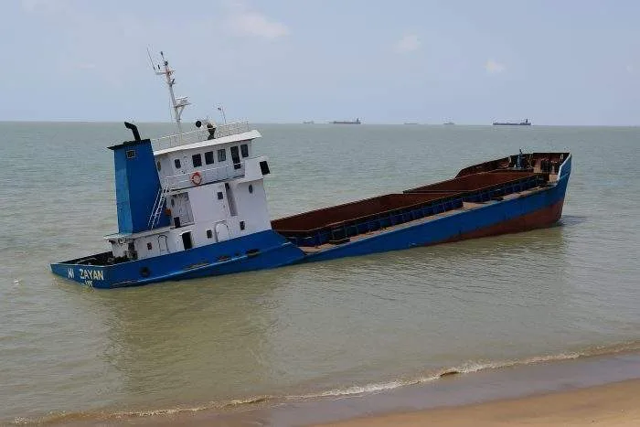 A partially sunken blue cargo ship named MV Zayan lies tilted near the shore at Chattogram Port, Bangladesh.