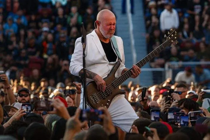 Limp Bizkit bassist performs passionately on stage in front of a large concert crowd, wearing a white suit and playing his bass guitar.