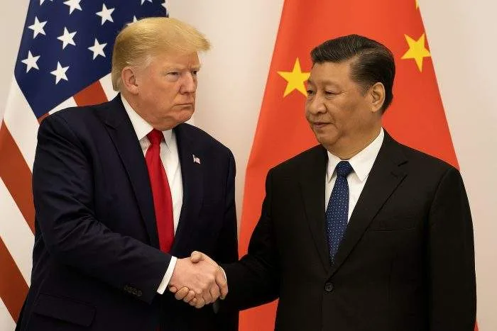 Donald Trump and Xi Jinping shake hands during a diplomatic meeting, standing before U.S. and Chinese flags.