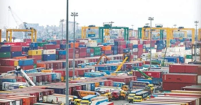 A busy view of Chattogram Port, Bangladesh, showing rows of colorful shipping containers, cranes, and cargo trucks under bright daylight.