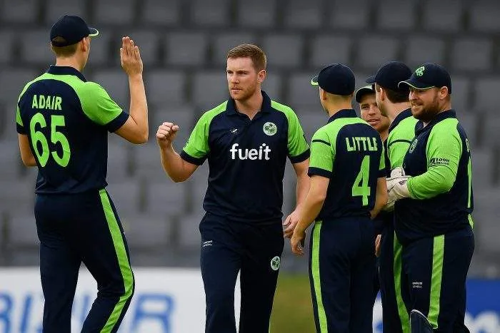 Ireland cricket players celebrate during a match, with fast bowlers Mark Adair and Josh Little seen in team huddle wearing green-and-navy jerseys.
