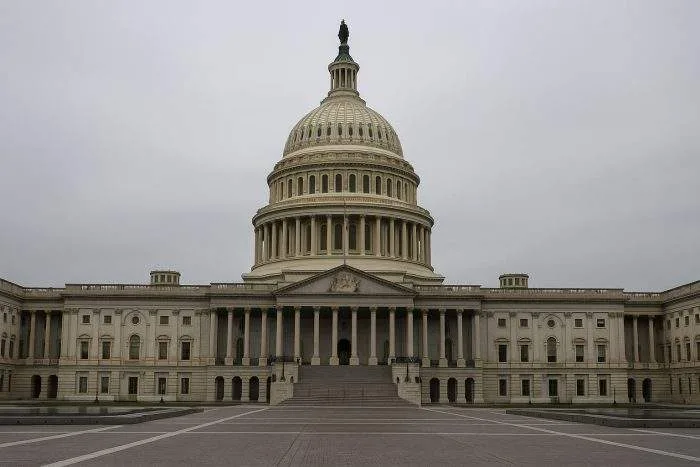 The U.S. Capitol building in Washington, D.C., stands under a gray sky with its dome and facade prominently visible, symbolizing political gridlock during a government shutdown.