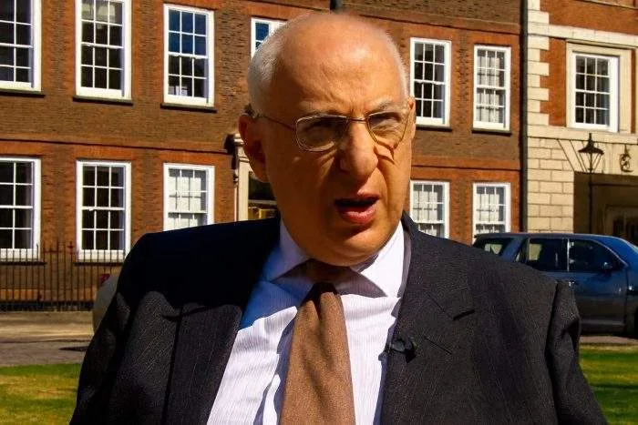 An older man in a suit and tie being interviewed outdoors in front of a historic red-brick building.