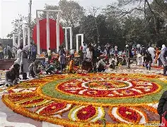 A horizontal wide-angle photo showing a group of university teachers in traditional white and black attire, standing barefoot on the steps of the Central Shaheed Minar, holding a large floral wreath with "Dhaka University Teachers’ Association" written on a ribbon.