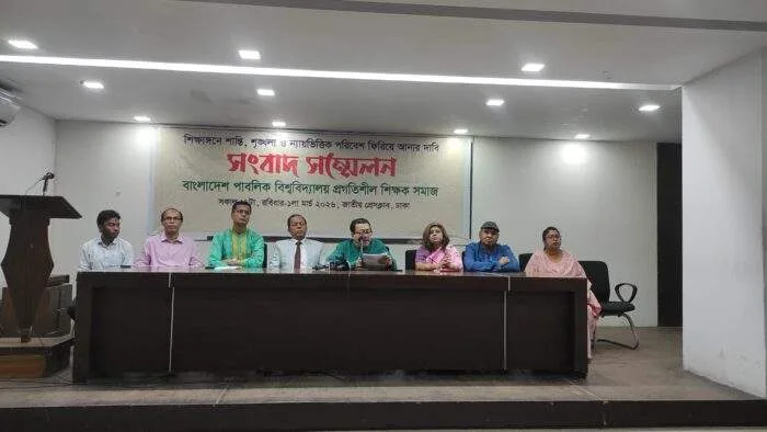 A group of university professors sitting behind a banner at the National Press Club in Dhaka, holding a press conference with multiple microphones in front of them.