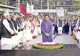 A wide shot of the "Swadhinata Stambha" (Independence Monument) at Suhrawardy Udyan in Dhaka, with scattered groups of citizens laying wreaths in a quiet, unofficial ceremony.