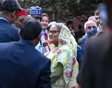A dignified portrait of Sheikh Hasina wearing a simple, off-white Shovon sari, seated in a library-like setting. Beside her is a framed photograph of Bangabandhu Sheikh Mujibur Rahman. The lighting is soft but firm, emphasizing a sense of "Unfinished Struggle."