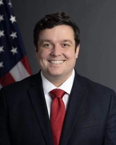 Official portrait of a U.S. State Department official standing in front of an American flag