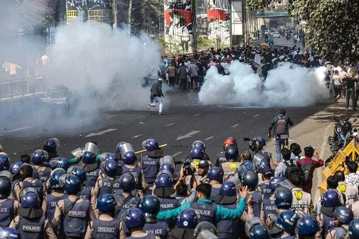 Police in riot gear face protesters on a Dhaka street as tear gas fills the air during a clash.