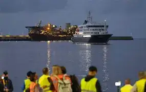 Passengers in white protective hazmat suits boarding a chartered evacuation flight at Tenerife North Airport, with the MS Hondius visible in the distance.