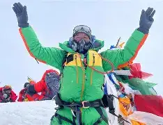 A long queue of mountaineers in colorful down suits stepping along a narrow, snowy ridge toward the summit of Mount Everest against a bright blue sky.
