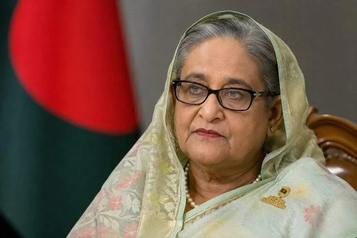 Former Bangladesh Prime Minister Sheikh Hasina in a formal portrait setting, photographed against a softly blurred Bangladesh flag backdrop.
