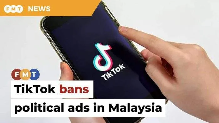 A close-up of a smartphone screen displaying the TikTok app logo, held against a blurred background of the Malaysian federal administrative capital or royal palace.
