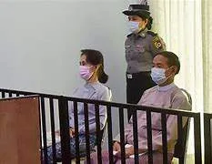 A split-screen editorial graphic showing a recent state-media image of Aung San Suu Kyi and a symbolic representation of a military-guarded residence in Nay Pyi Taw.