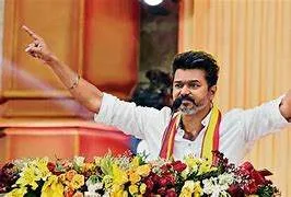 Actor and politician Vijay standing on a podium, addressing a massive, cheering crowd under the banner of Tamilaga Vettri Kazhagam.