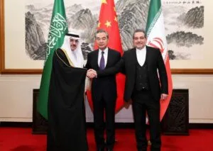 A high-resolution horizontal photograph of a bilateral diplomatic meeting room. Saudi Foreign Minister Prince Faisal bin Farhan and Chinese Foreign Minister Wang Yi stand side-by-side on a red carpet, shaking hands professionally. Behind them are the national flags of Saudi Arabia and the People's Republic of China, capturing a major geopolitical moment of Middle Eastern and Asian diplomacy.