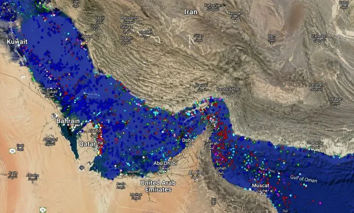 A high-resolution horizontal photograph of a large commercial oil tanker navigating through a narrow, sunlit maritime strait bounded by rugged, arid coastal mountains in the background. The water shows slight white wakes from the vessel's movement, capturing a tense and highly strategic global energy chokepoint.