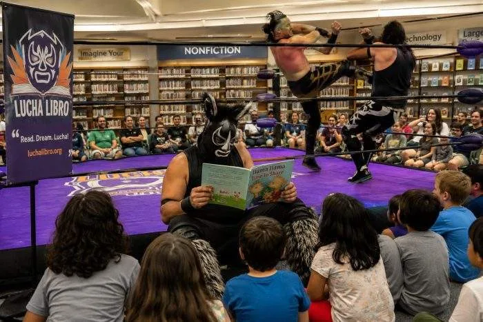 Horizontal view of a professional wrestling ring set up inside a public library surrounded by bookshelves.