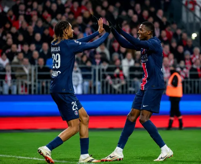 France forward Ousmane Dembélé holding up three fingers in celebration on the pitch at Boston Stadium. He is wearing the light blue French national team away kit with the iconic golden cockerel crest visible. A teammate runs up behind him smiling, while a stadium crowd fills out the background under bright daylight.