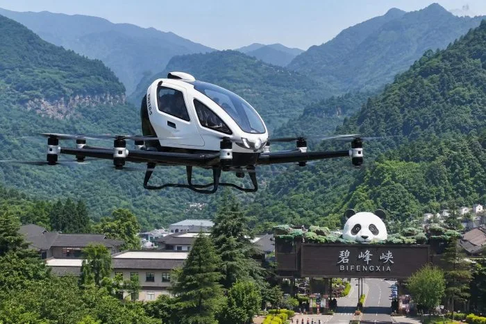 A sleek, modern electric vertical take-off and landing (eVTOL) aircraft with multiple rotors gliding smoothly over a deep, lush green river canyon in Sichuan. The sky is partially filled with low-lying clouds drifting between the mountain peaks. The aircraft is captured mid-flight, highlighting its clean aerodynamic lines and futuristic design.
