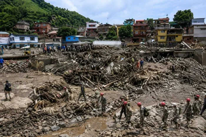 A serious editorial photograph of a professional rescue team wearing helmets and high-visibility vests working amidst the concrete rubble of a partially collapsed building structure. Emergency service vehicles with flashing lights are visible in the blurred background under a hazy sky, capturing the intense and coordinated disaster response effort.