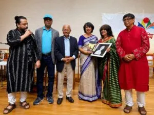 A dignified horizontal photograph capturing author Nurjahan Bose standing proudly on an indoor event stage, holding a large, golden-framed Lifetime Achievement Award. She is dressed in an elegant patterned sari. Beside her, an organizer speaks into a microphone, while a sitting audience applauds warmly. The stage background features large event banners for the Bangla Center Book Fair.