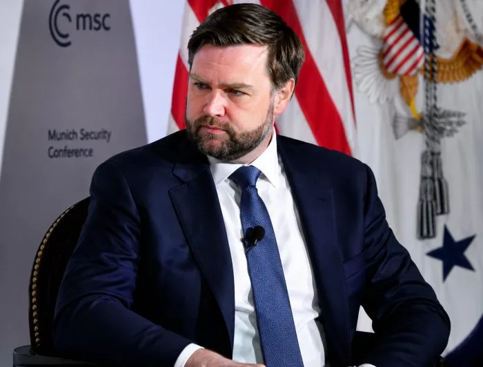 A professional editorial photograph capturing U.S. Vice President JD Vance speaking from a microphoned podium inside a formal Washington briefing room. The background features a softly blurred American flag, accentuating the high-stakes and serious nature of the geopolitical announcement.