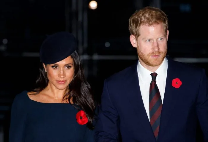 A professional editorial photograph of Prince Harry and Meghan Markle standing together at a formal evening event. Prince Harry is dressed in a classic dark suit, while Meghan Markle wears an elegant, neutral-toned formal dress. They are smiling warmly, captured in a clean, journalistic style with a softly blurred background.