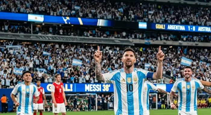 A crisp, wide-angle sports photograph capturing Lionel Messi celebrating his historic goal under stadium floodlights. He is pointing both hands toward the sky with a triumphant expression, while teammates in Argentina's iconic blue and white striped shirts rush toward him to celebrate. The packed World Cup stadium blurred in the background highlights the intense energy of the moment.