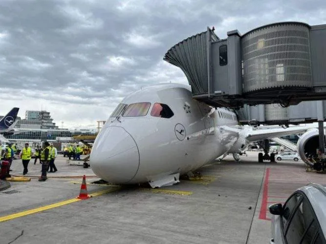 A large commercial Boeing 787-9 passenger jet resting awkwardly on its nose on the airport tarmac, surrounded by flashing emergency response vehicles and ground crew.