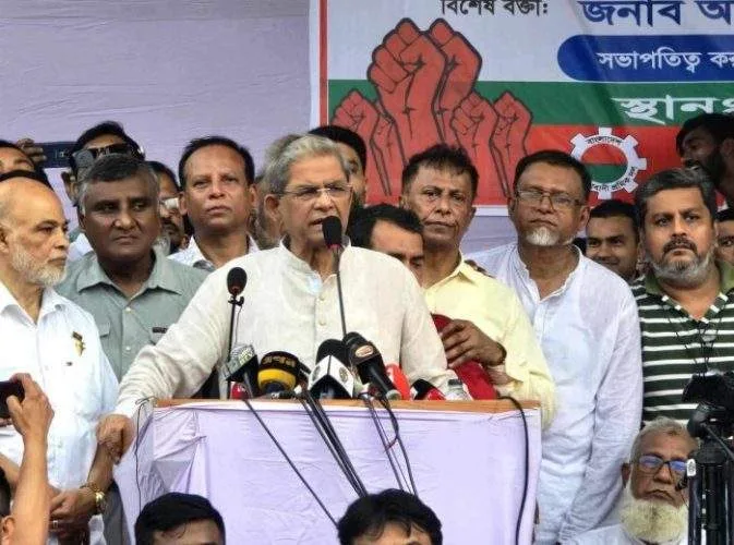 BNP Secretary General and LGRD Minister Mirza Fakhrul Islam Alamgir standing at a decorated podium, addressing a massive public gathering at Kumarpur High School grounds.