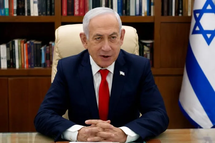 A professional editorial photograph of Israeli Prime Minister Benjamin Netanyahu speaking at a press conference podium. Microphones are clustered in front of him, and the blue and white flag of Israel stands prominently in the background, capturing a high-stakes national security announcement.