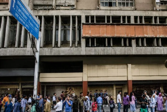 A close-up photograph of the metal security shutter pulled entirely down over a main entrance to a metro station in Caracas. A printed official notice paper is taped to the shutter, reading "SERVICIO SUSPENDIDO" (Service Suspended) in bold lettering. In the blurred background, empty and unusually quiet city streets stretch out under a hazy sky, capturing the immediate nationwide halt of public activities.