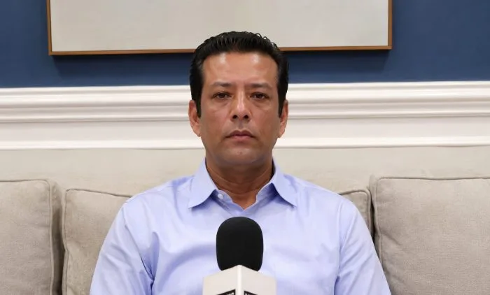 A professional file portrait photograph of Sajeeb Wazed Joy speaking seriously, wearing a dark formal vest over a light-colored shirt. The background is softly blurred to place full focus on his expression, capturing an authoritative and high-profile political commentary tone.