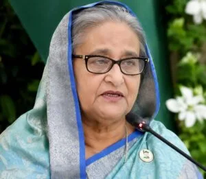 A professional file or broadcast portrait photograph of former Prime Minister Sheikh Hasina during an official interview. She is wearing a traditional sari, maintaining a somber and determined expression as she speaks. The background is softly blurred to focus entirely on her face, capturing a high-stakes and formal political presentation.