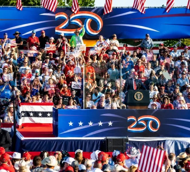 A wide-angle, realistic photograph of the National Mall in Washington, D.C., decorated with large American flags and grand event staging under a clear blue sky. Crowds of spectators gather on the lawns facing the Washington Monument, with military jets visible in a precise formation overhead, capturing a momentous national anniversary celebration.