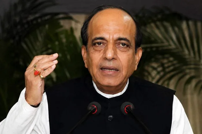 A professional medium-shot photograph of India's newly appointed High Commissioner to Bangladesh, Dinesh Trivedi, at a formal state event in Dhaka. He is wearing a formal Nehru jacket suit and sharing a handshake with a senior official. The background features the national flags of India and Bangladesh placed side-by-side in an elegant, well-lit ceremonial hall, capturing a dignified and authentic diplomatic atmosphere.