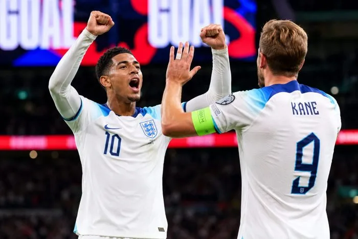 England captain Harry Kane in his white national jersey shouting in celebration on the pitch. His golden captain's armband is visible on his arm, and the crowded stands of the stadium fill the background under a modern canopy roof.