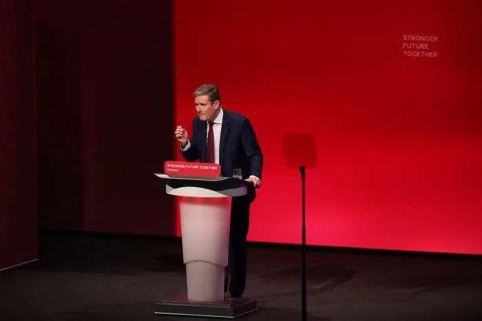 British Prime Minister Keir Starmer speaking with a stern expression behind a dark wood podium displaying the UK government emblem.