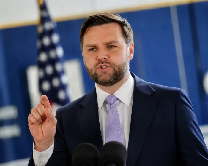 A high-resolution horizontal photograph of U.S. Vice President JD Vance speaking during an interview on a television news set. He is wearing a dark suit with a blue tie, gesturing with his hand as he explains the administration's strategic position regarding diplomatic frameworks, with a studio backdrop displaying global maps.