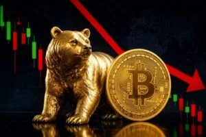 A professional, close-up photograph of a golden physical Bitcoin coin resting on a dark, reflective surface with red and green digital stock market candlestick charts softly blurred in the background.