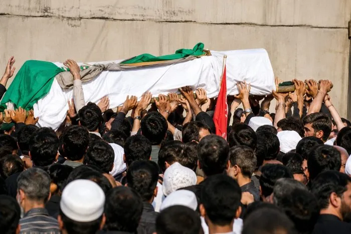 A wide-angle, horizontal photograph showing the massive exterior of Tehran's Grand Mosalla Mosque, surrounded by immense crowds of mourners gathering under a somber, overcast sky with heightened security forces stationed at checkpoints.