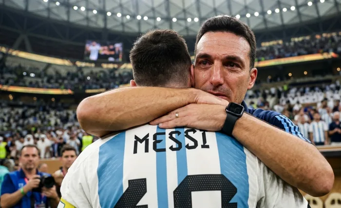 A tearful Argentina manager Lionel Scaloni sharing an emotional hug with captain Lionel Messi from behind, showcasing the iconic number 10 jersey in a packed stadium during their historic World Cup victory celebration.