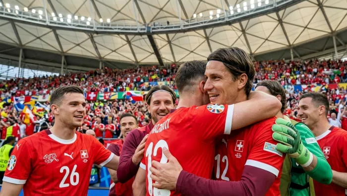 A joyful close-up photograph capturing a group of Swiss national football players celebrating. The central player with a black headband is hugging another player from behind, smiling widely. Their red kits, with the 'SUISSE' crest and number 12, are clear, as is a prominent green goalkeeper's glove. The background is a massive stadium crowd and its unique dome roof structure.